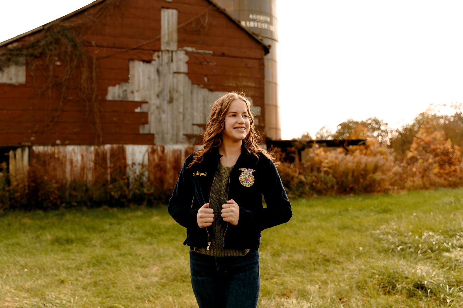 Young woman in FFA jacket smiling outdoors near a rustic red barn on a farm.