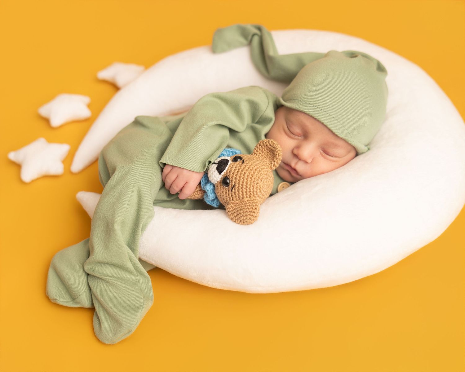 Newborn baby sleeping peacefully in green outfit and hat, cuddling a knit bear toy on white moon pillow.