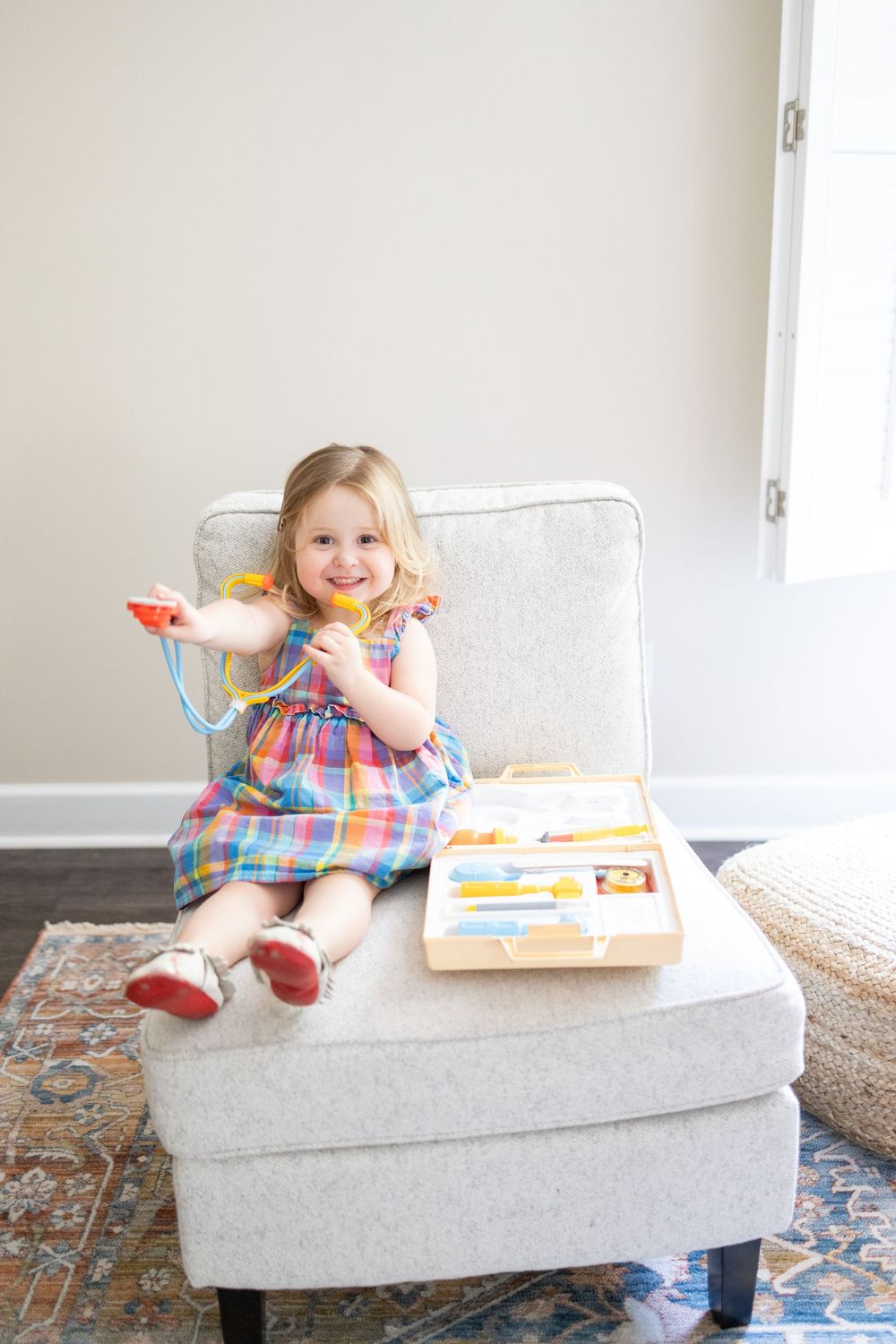 A cheerful toddler girl in a plaid dress plays with a toy doctor kit on a gray chair.