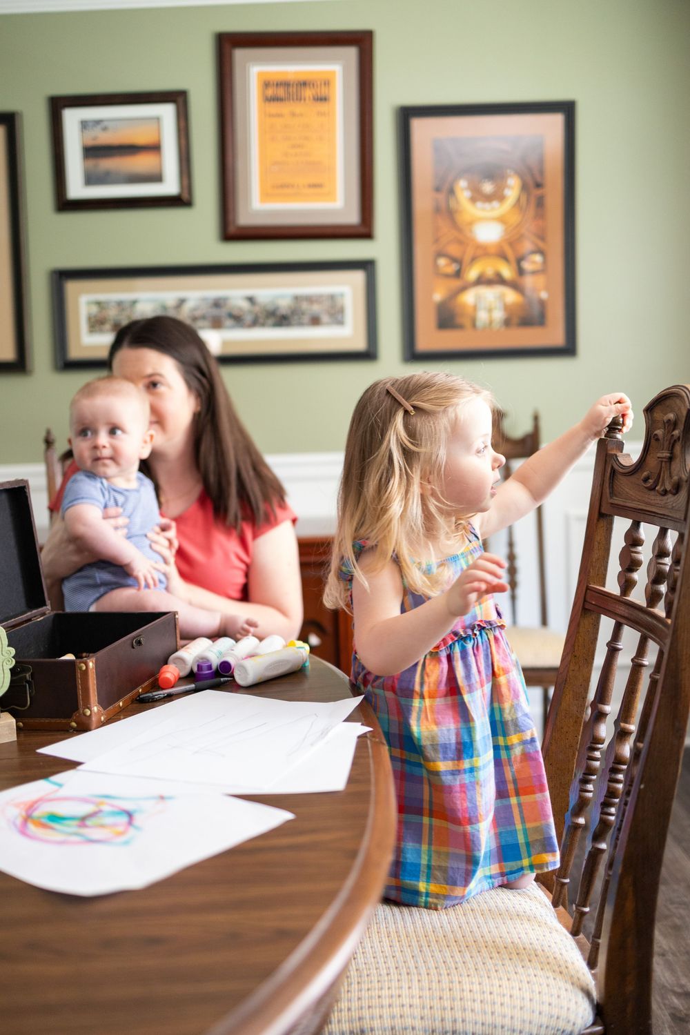 Mother holding baby at home office desk while young girl in plaid dress plays nearby with framed art on wall.