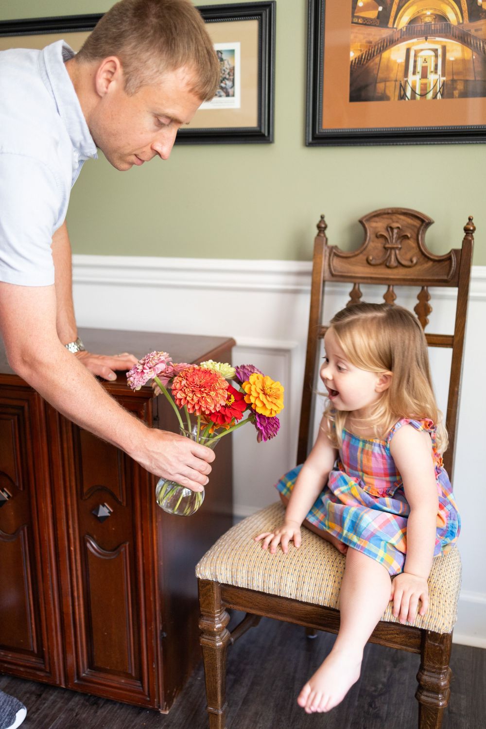 Father showing colorful flower bouquet in glass vase to smiling toddler girl sitting on wooden chair indoors.