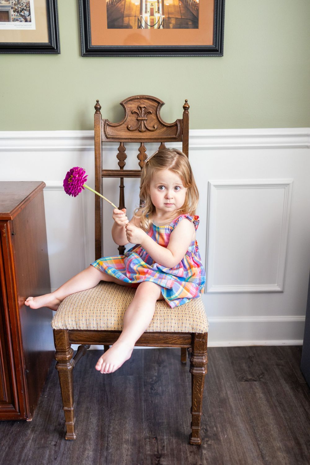 Young girl in colorful dress sitting on wooden chair holding pink flower indoors.