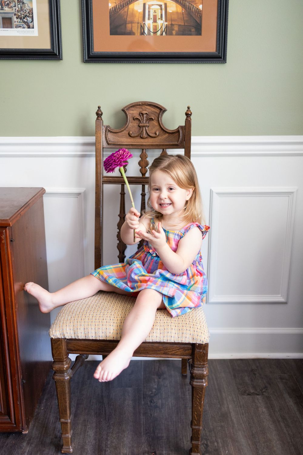 A smiling toddler girl in a colorful dress sits barefoot on a wooden chair, holding a pink flower.