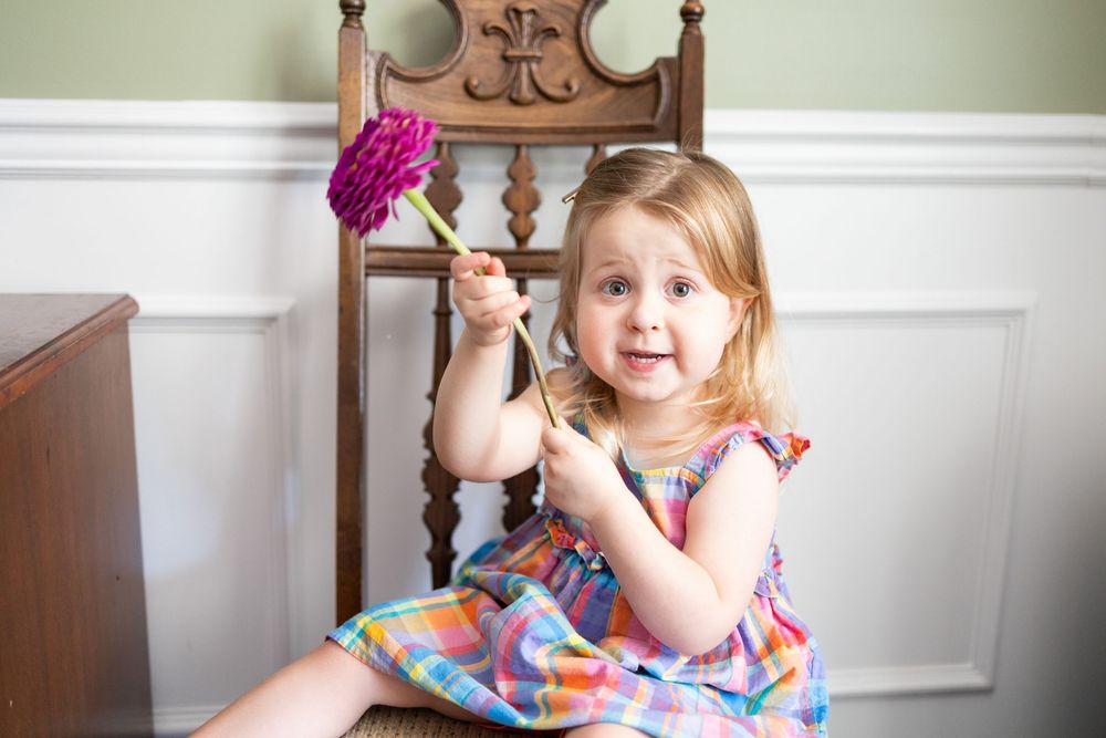 Cute toddler girl in colorful dress sitting on wooden chair, holding a pink feather duster toy indoors.