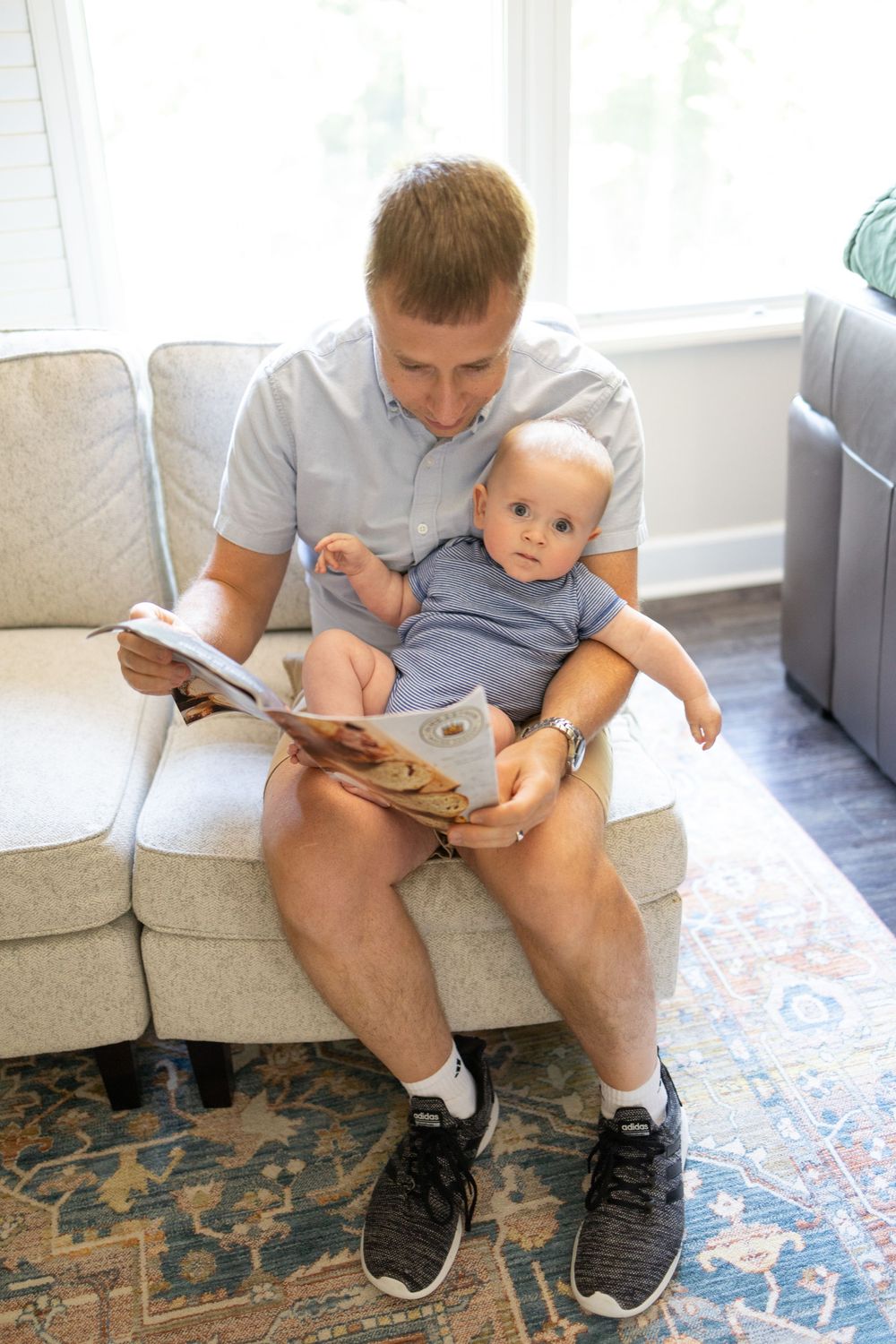 Father sits on couch reading King Arthur Baking cookbook while holding infant baby on his lap.