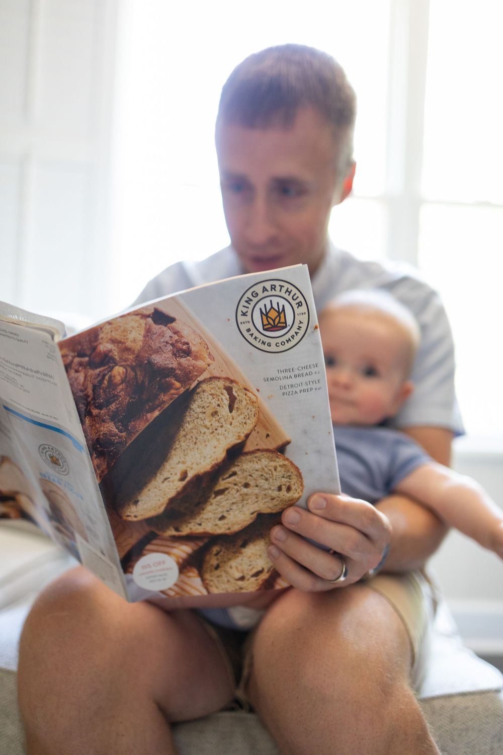 Close-up of King Arthur Baking Company cookbook held by father with baby on couch.