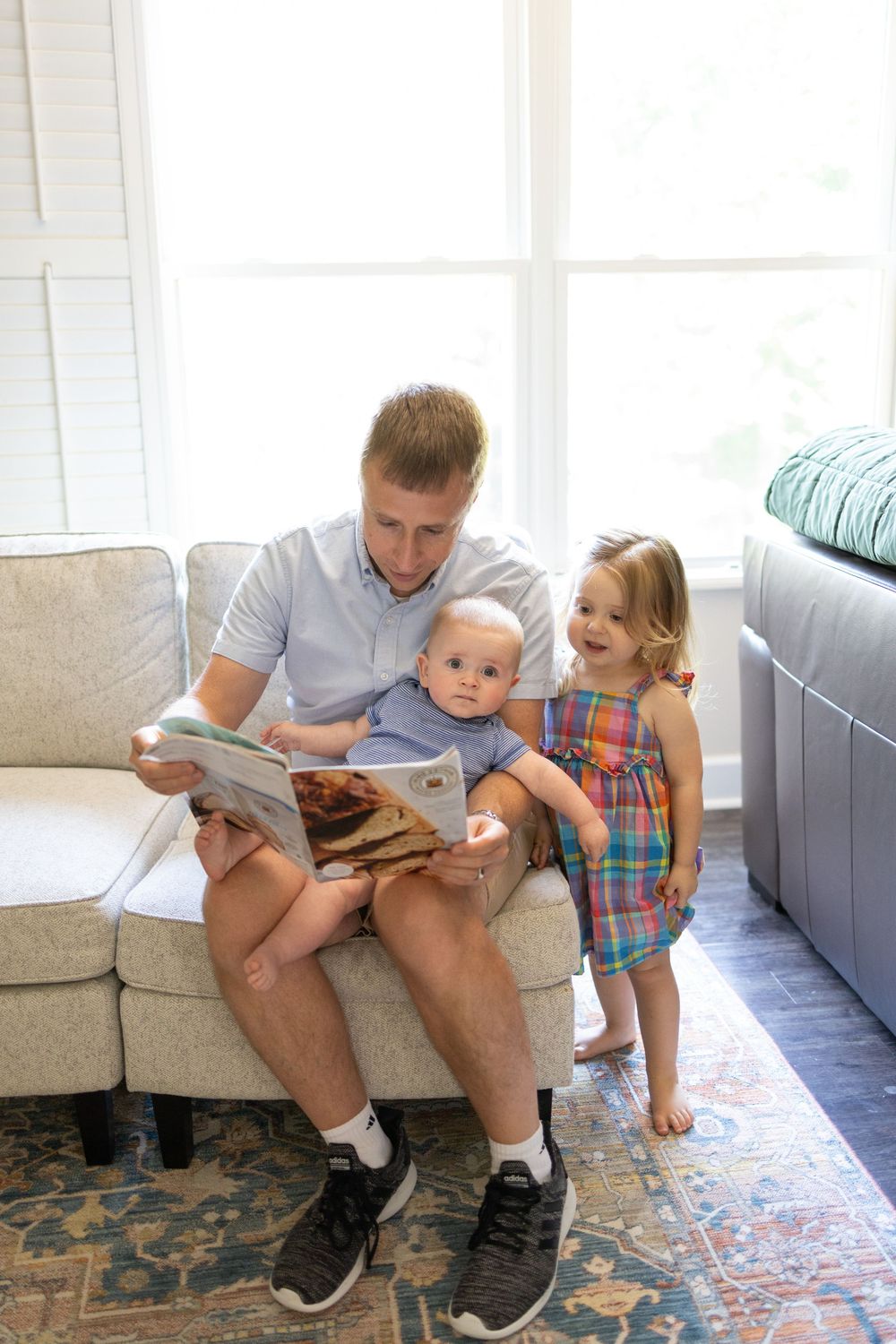 Father reading book to baby and toddler girl in plaid dress on light gray sofa by bright window.