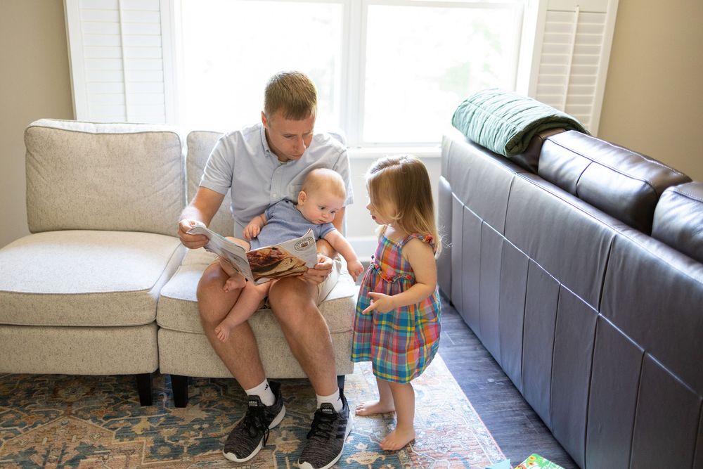 Father reads a picture book to his toddler girl and baby on a white couch in a bright living room.