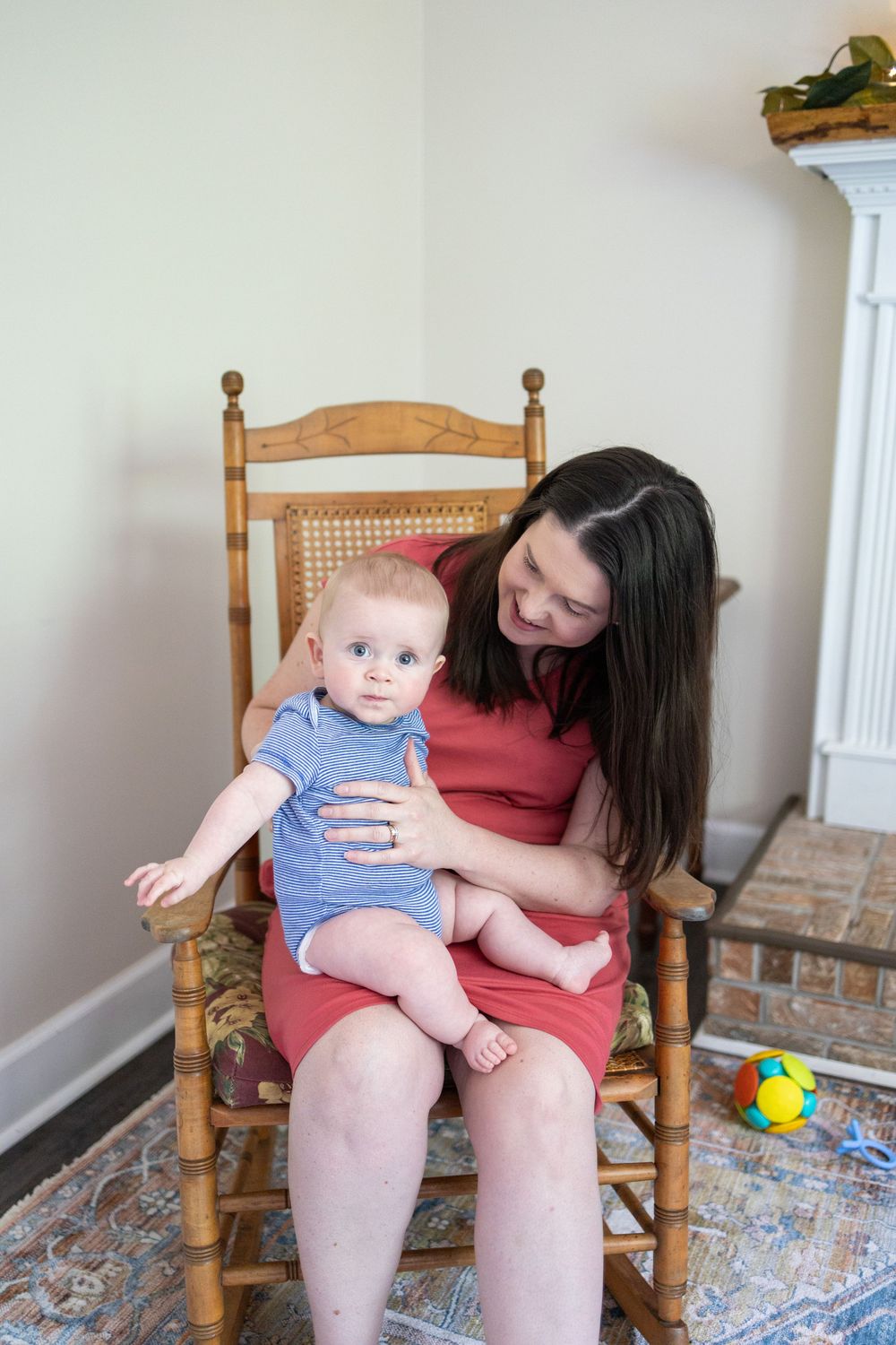 A smiling mother in a red dress holds her baby boy in a wooden rocking chair in a bright, cozy room.