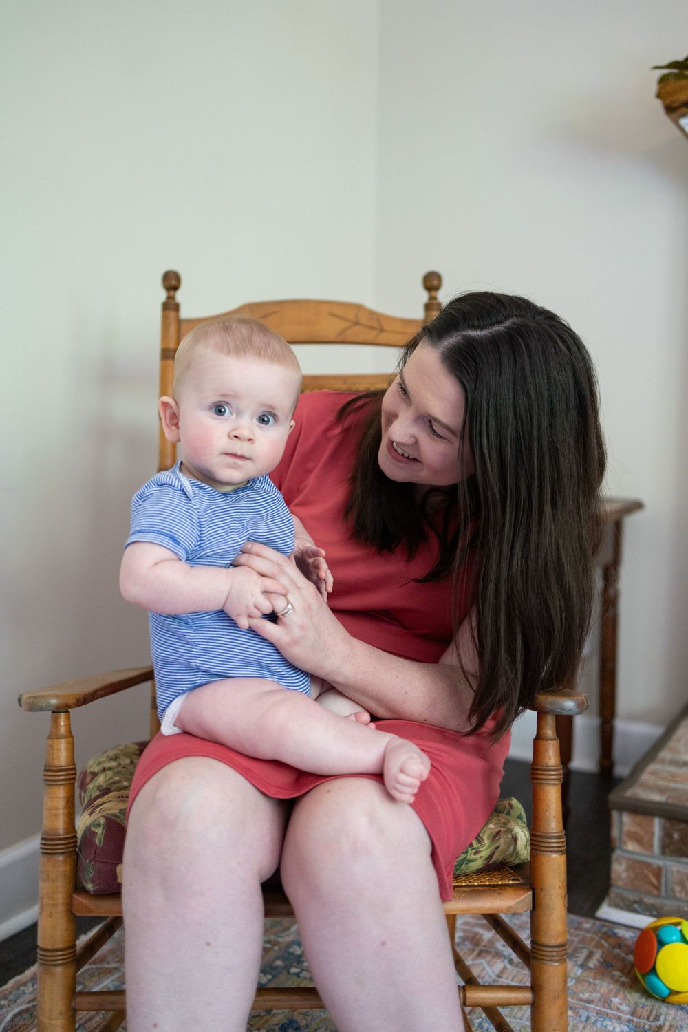 A smiling mother in a red dress holds her blue-eyed baby boy in a wooden rocking chair indoors.