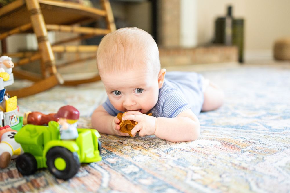 Baby in blue onesie lying on a rug, chewing a toy next to a colorful plastic tractor in a bright living room.