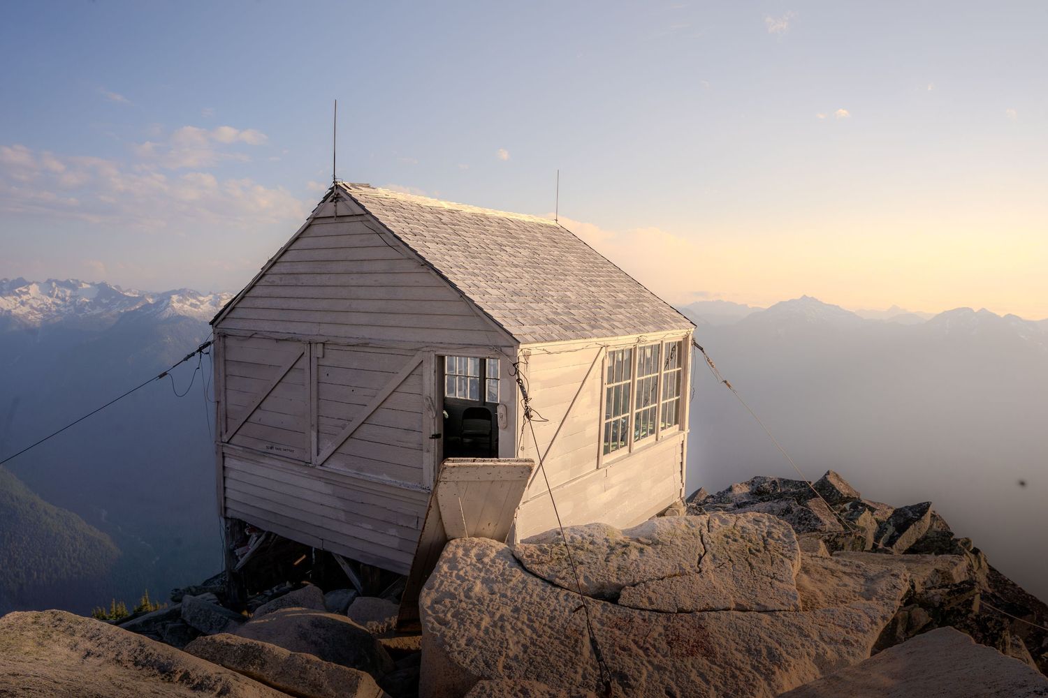 A rustic white wooden fire lookout cabin perched on rocky mountain summit at sunset with misty mountain views.