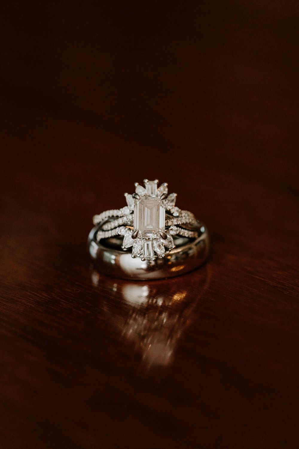 Elegant emerald cut diamond engagement ring with art deco halo stacked with wedding bands on dark wood surface.