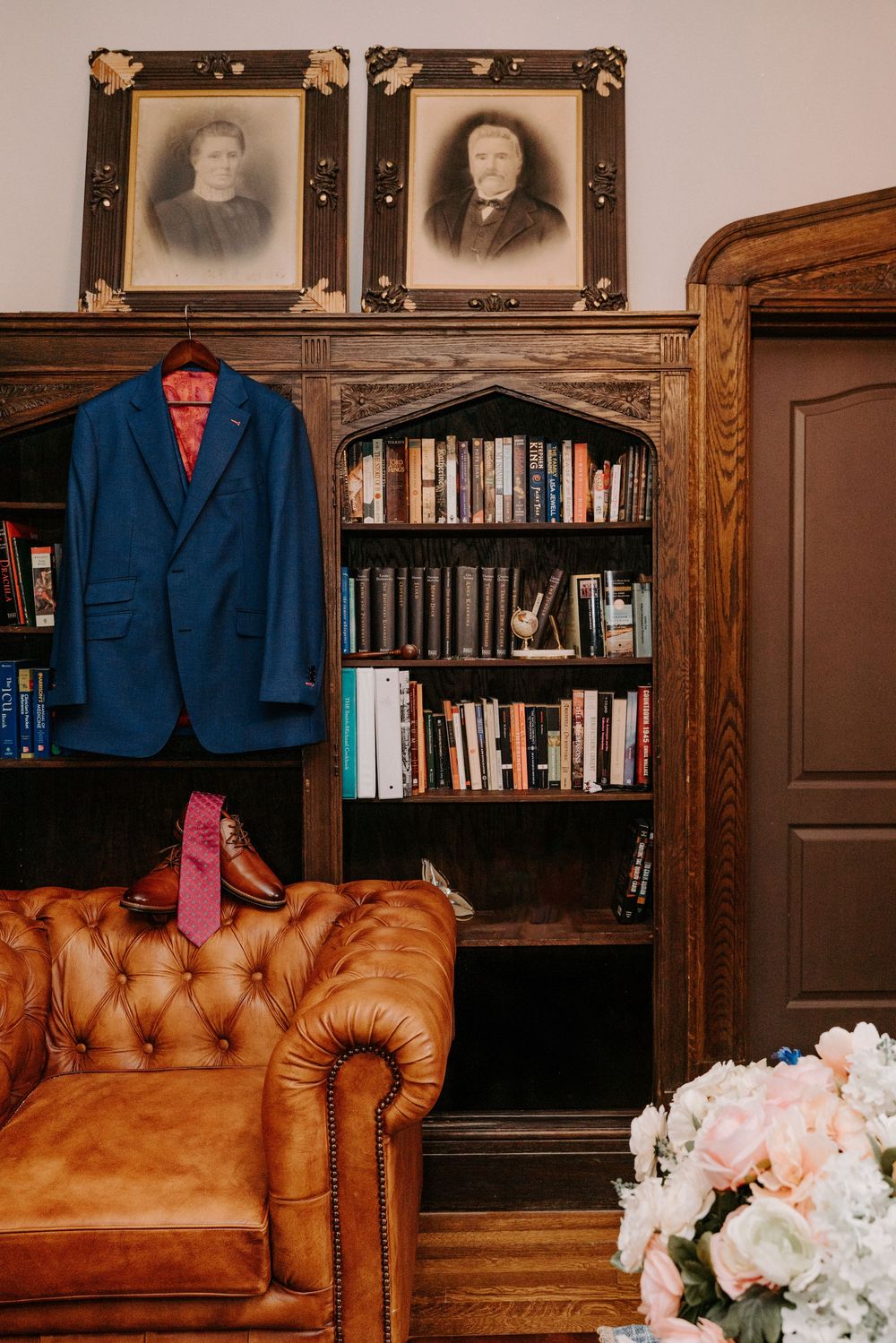 Blue suit jacket hanging in a wood-paneled library with leather Chesterfield sofa, books, and vintage portraits.