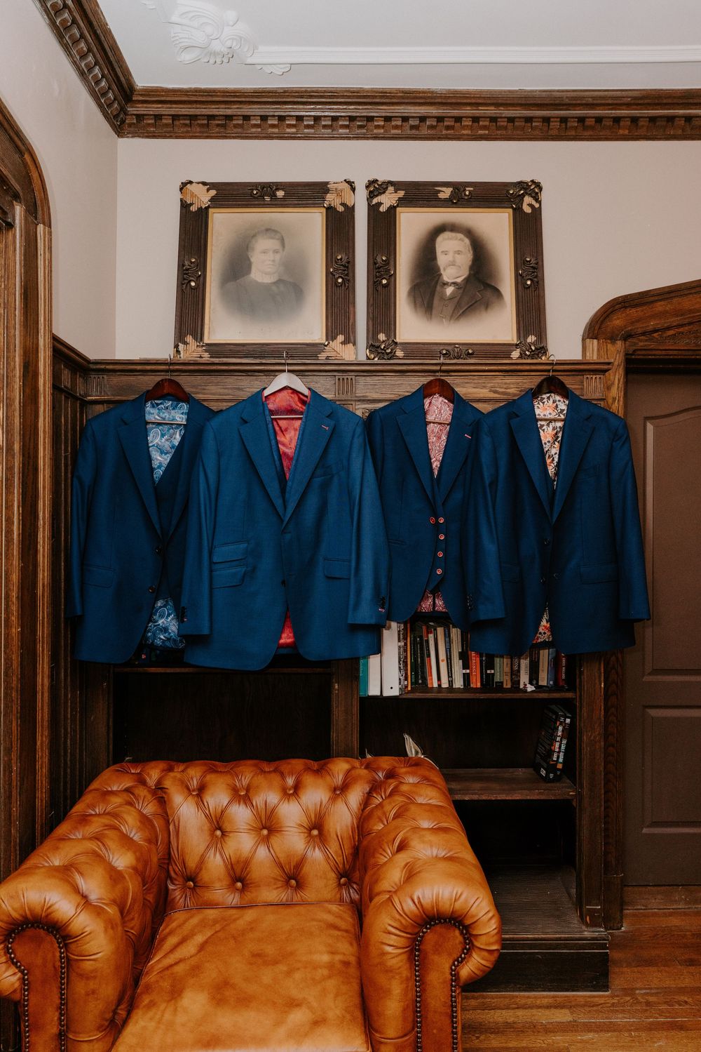 Four navy blue suits with colorful linings hang in a wood-paneled room with vintage portraits and a leather Chesterfield sofa.