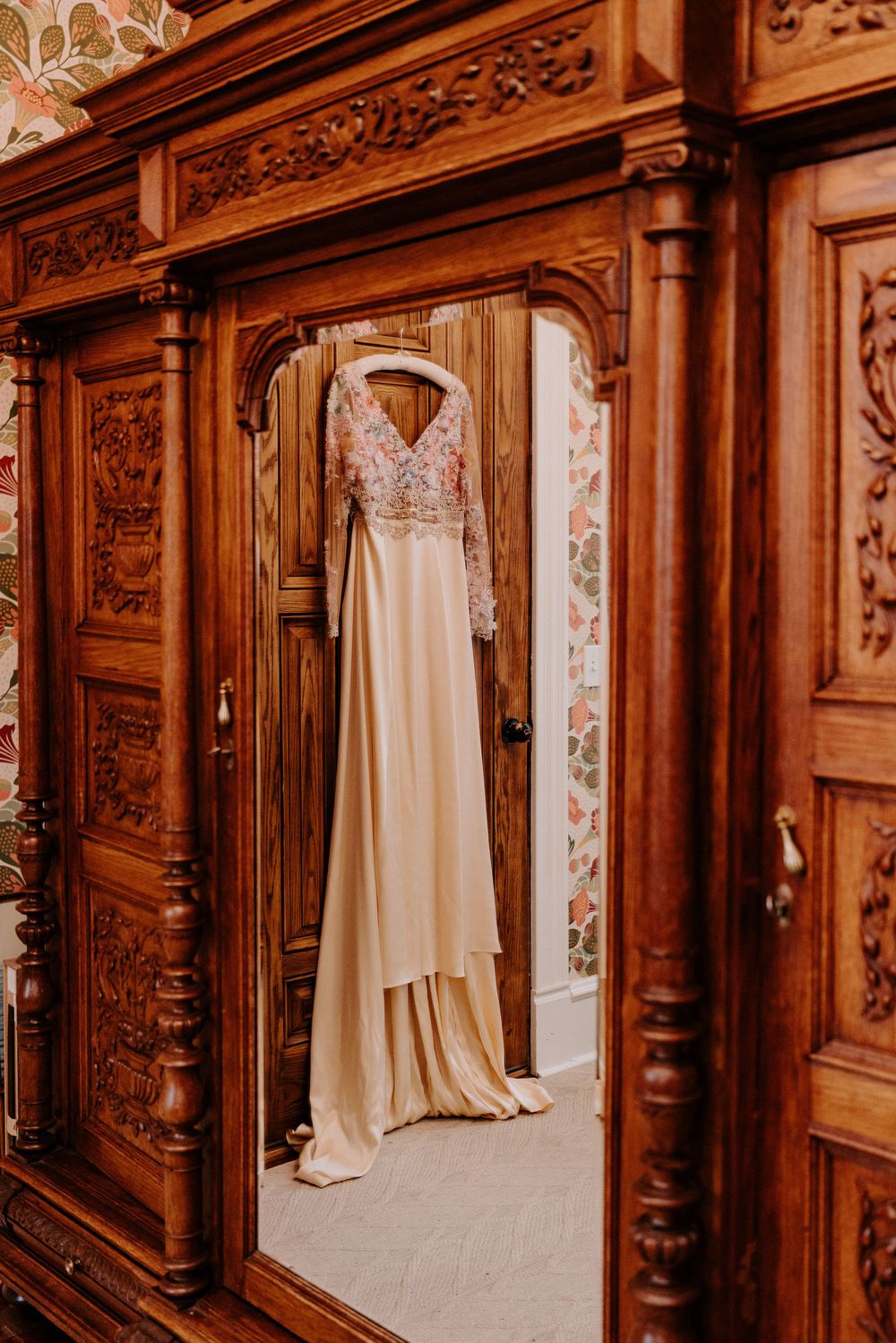 Elegant champagne wedding dress with floral bodice hanging in ornate carved wooden wardrobe mirror.