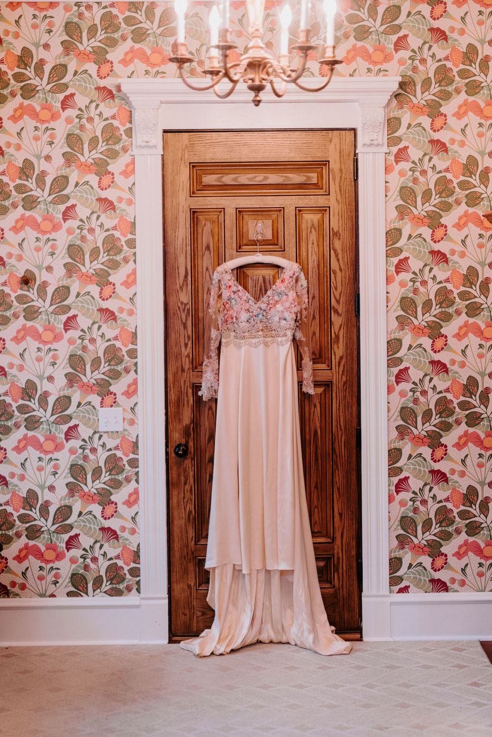 Blush wedding dress with floral embroidery hanging on wooden door with vintage floral wallpaper and chandelier.