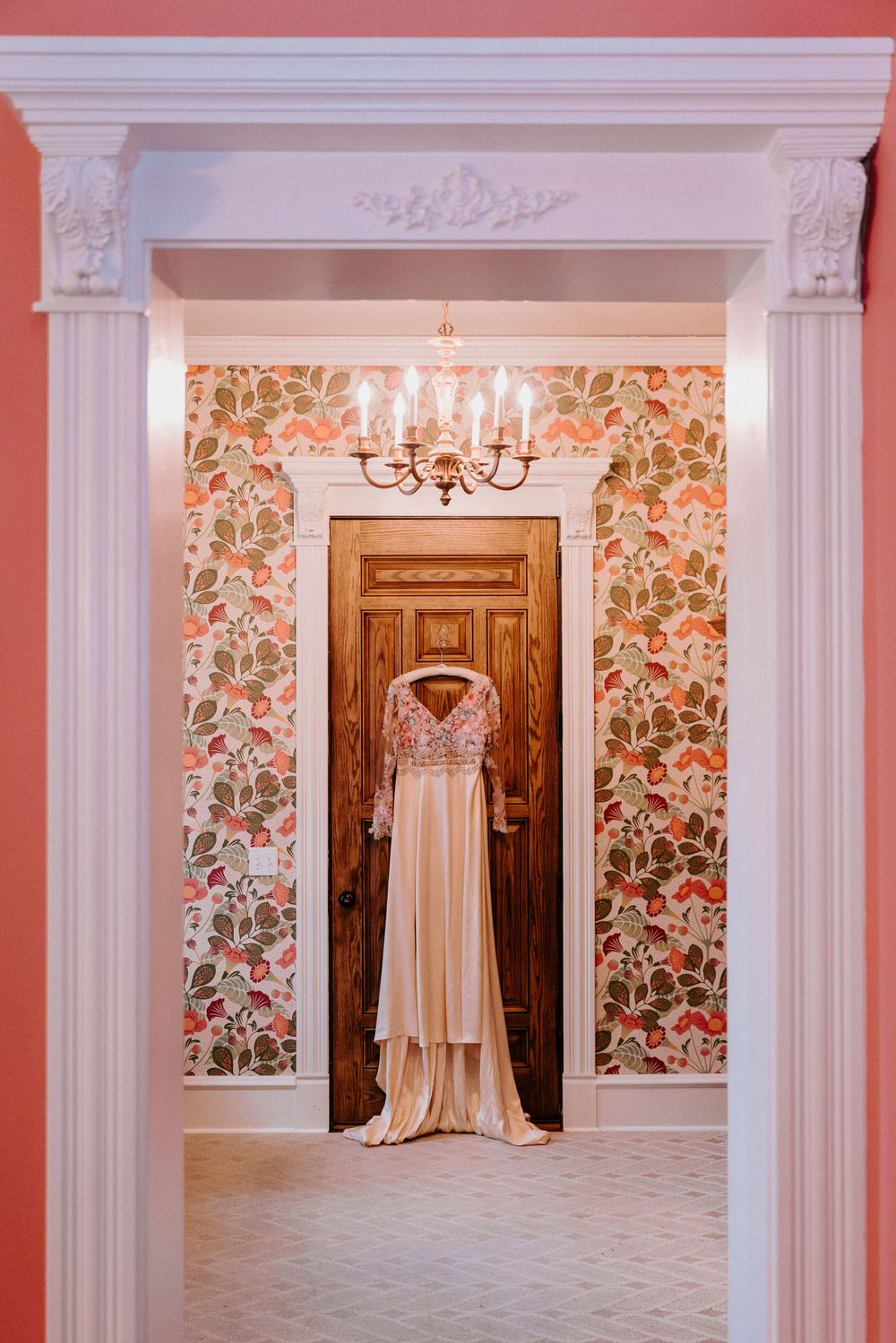 Elegant blush bridal gown displayed in doorway with ornate white molding, floral wallpaper, and chandelier.