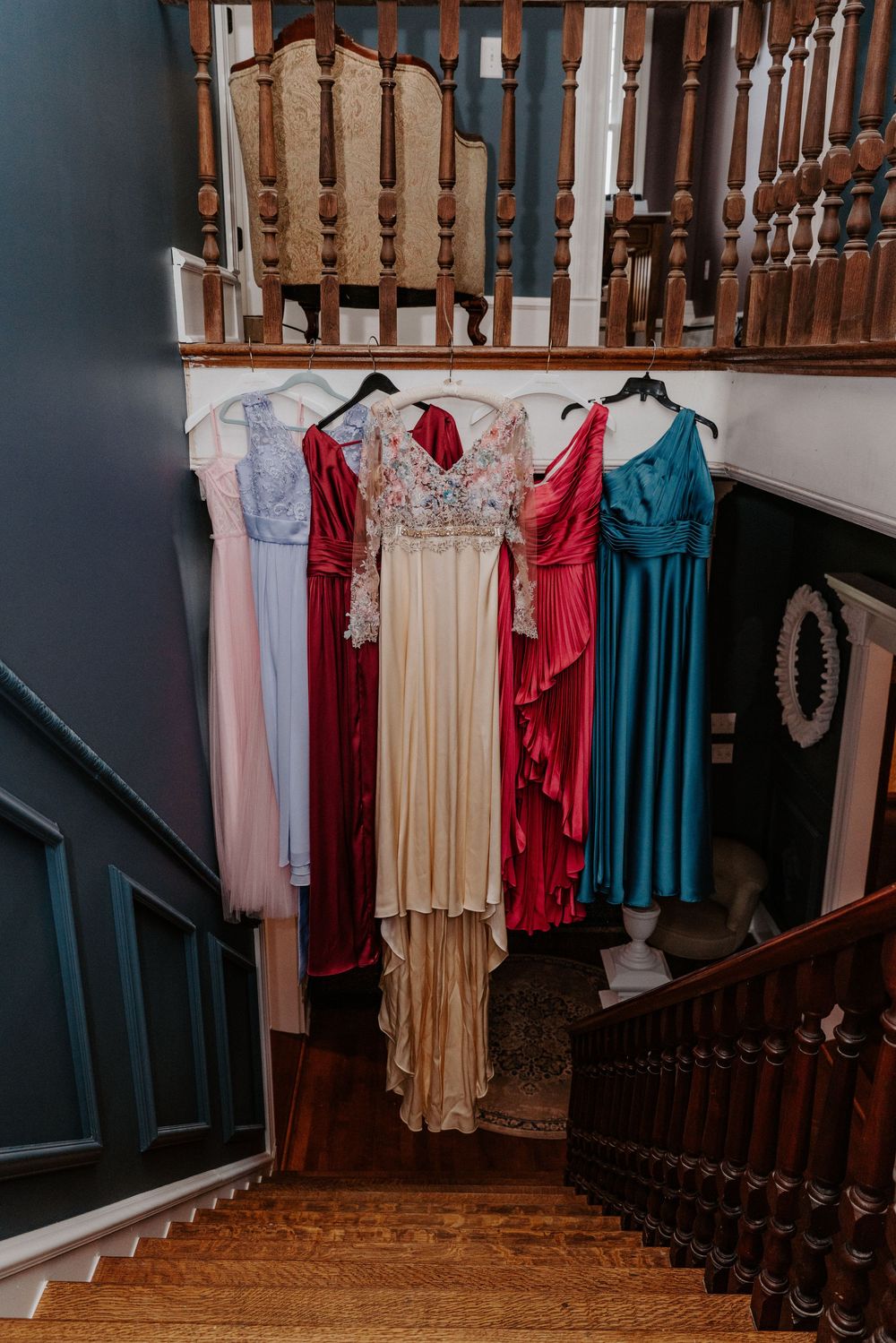 Five elegant formal gowns in pink, red, gold, and teal hanging on a wooden staircase banister in a classic home.