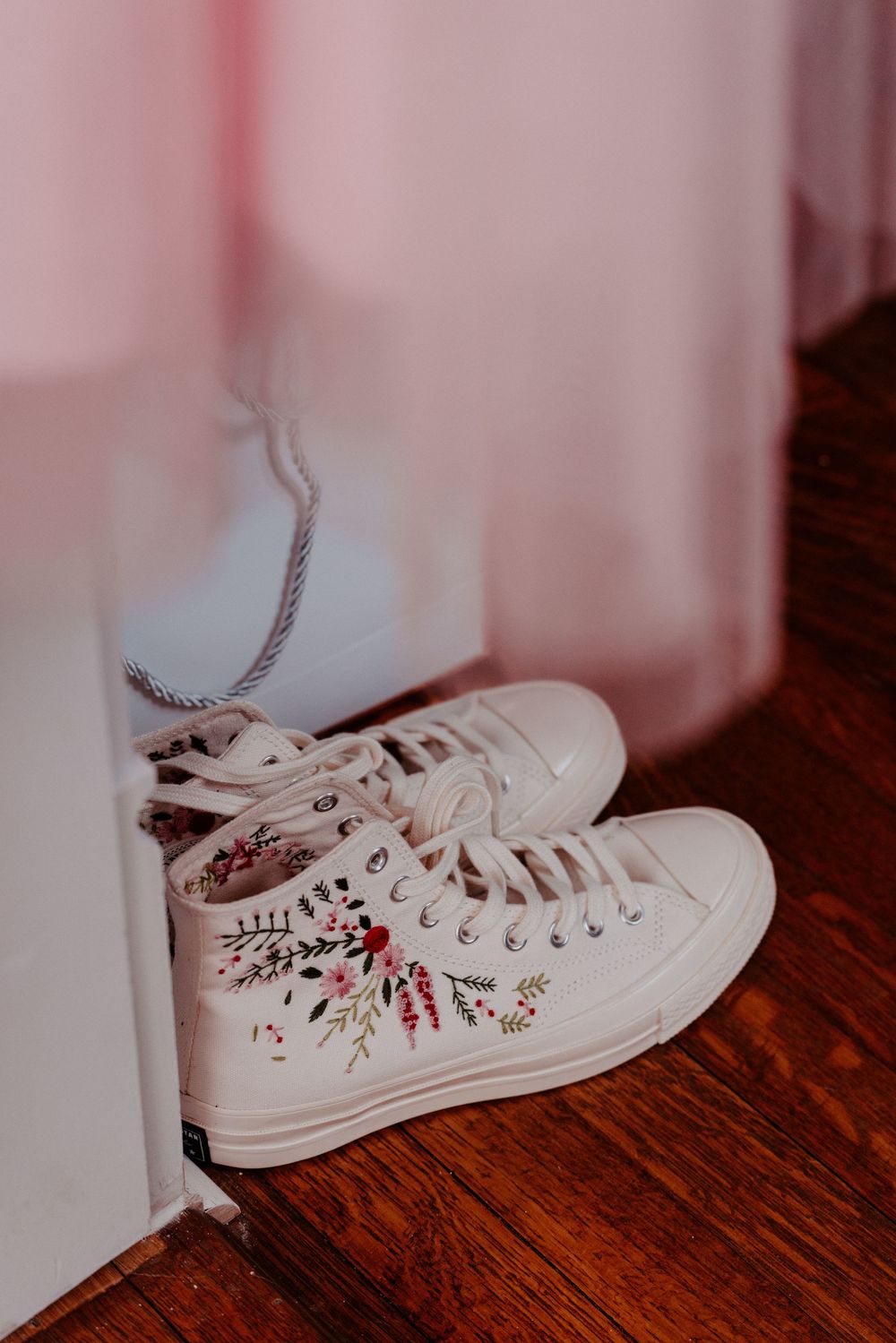 White high-top Converse sneakers with floral embroidery on hardwood floor near pink curtains.
