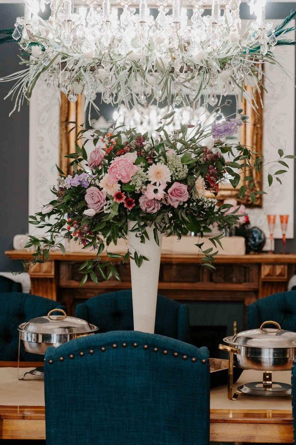 Elegant wedding reception table with floral centerpiece, teal chairs, chafing dishes, and crystal chandelier overhead.