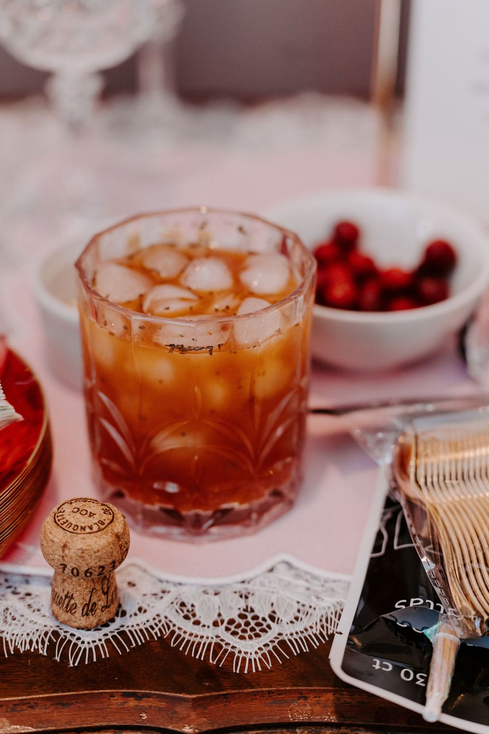 A vintage crystal glass with an amber cocktail on ice, beside a champagne cork and bowl of cherries.