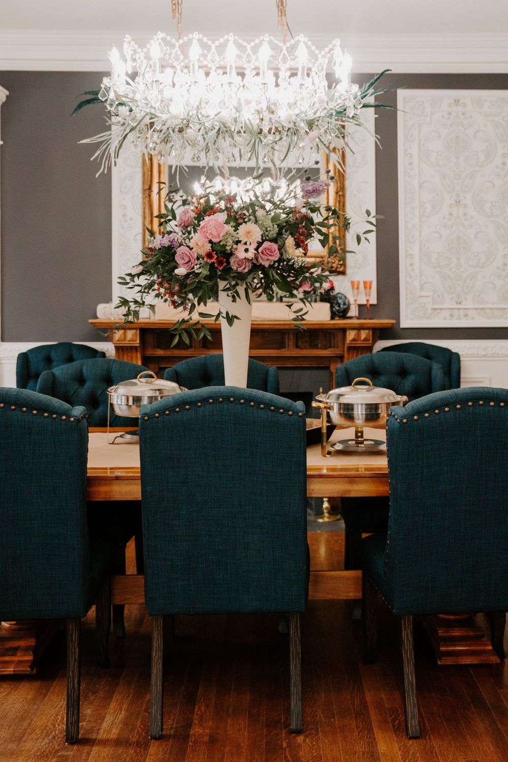 Elegant dining room with tufted teal chairs, wooden table, crystal chandelier, and pink floral centerpiece.