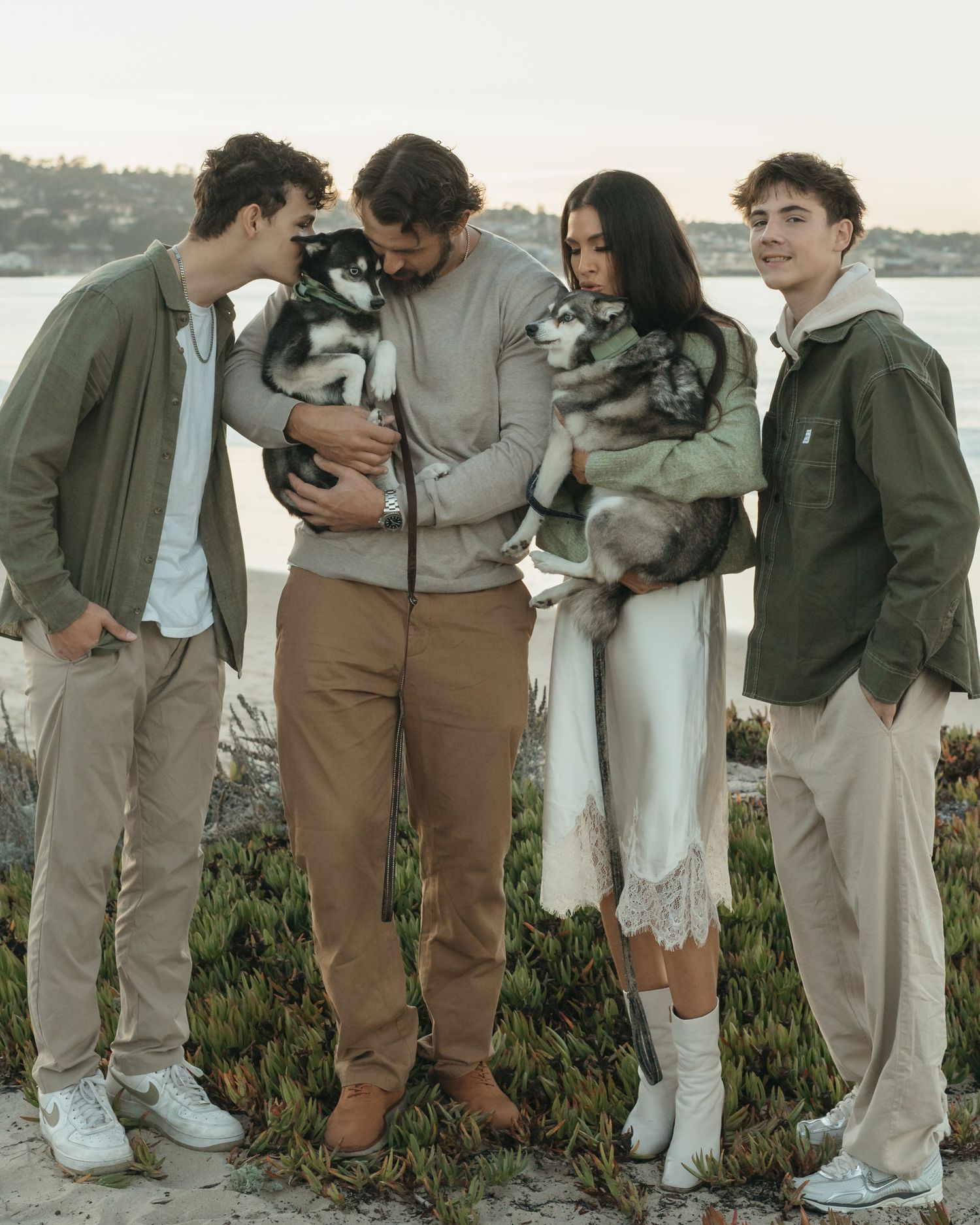 Four people holding two husky puppies outdoors near water, dressed in neutral tones for a casual family portrait.