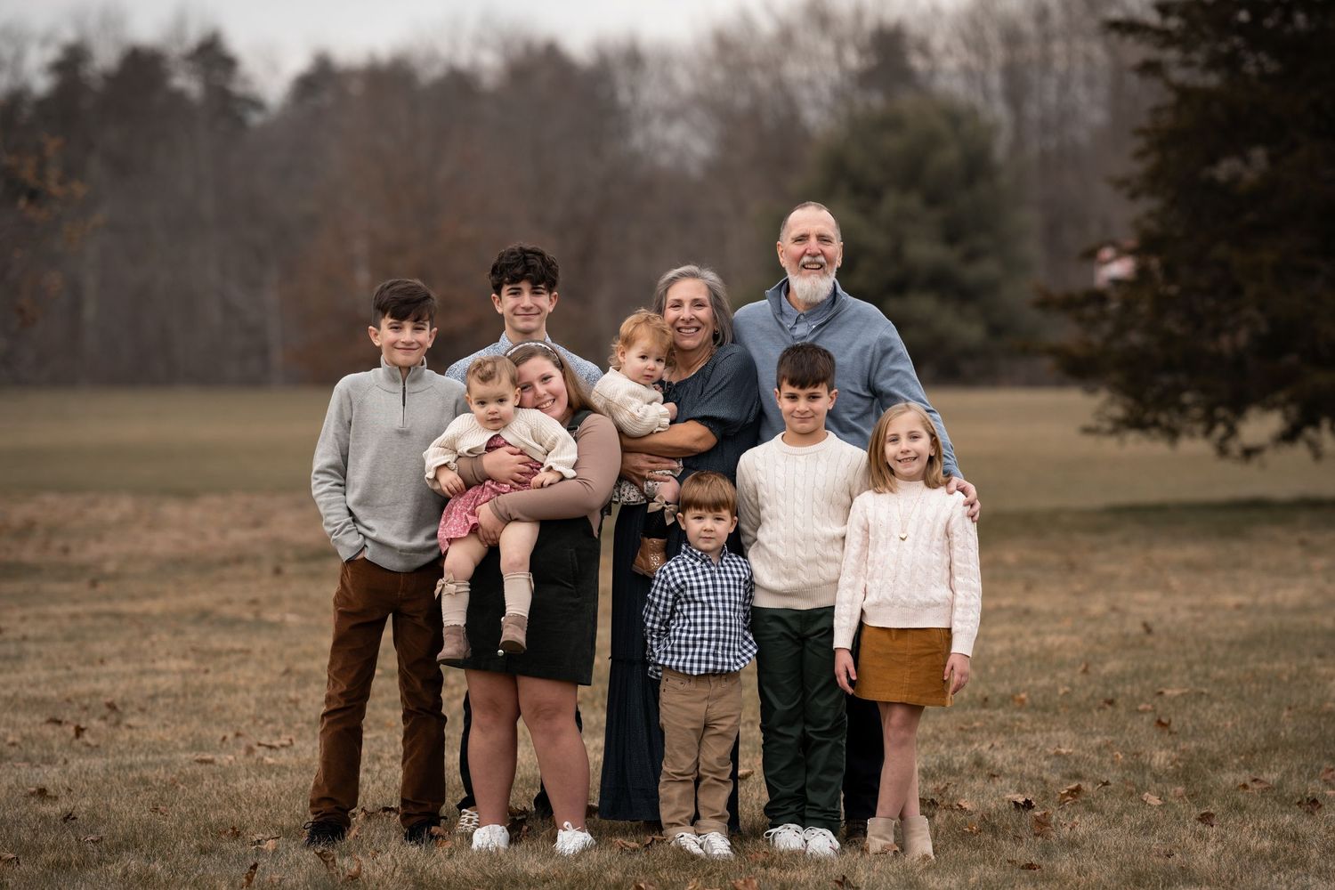 A group of seven people and a baby stand together outdoors in a field with trees in the background at dusk.