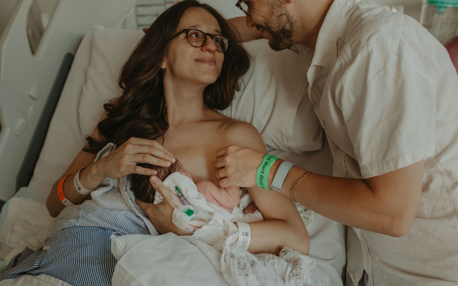 New parents sharing a tender moment in hospital bed while mother breastfeeds newborn baby after birth.