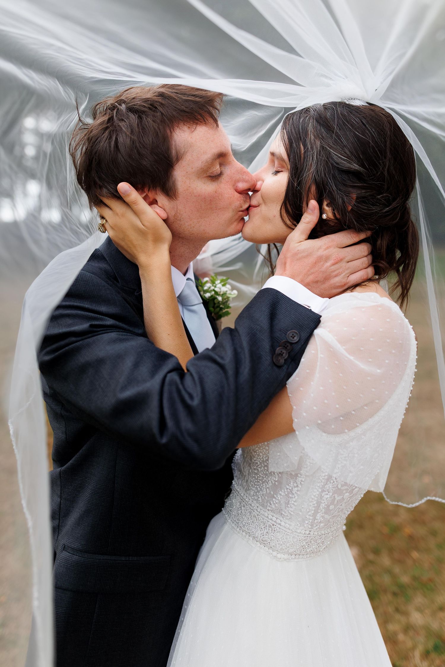 Bride and groom sharing a romantic kiss under a wedding veil outdoors.
