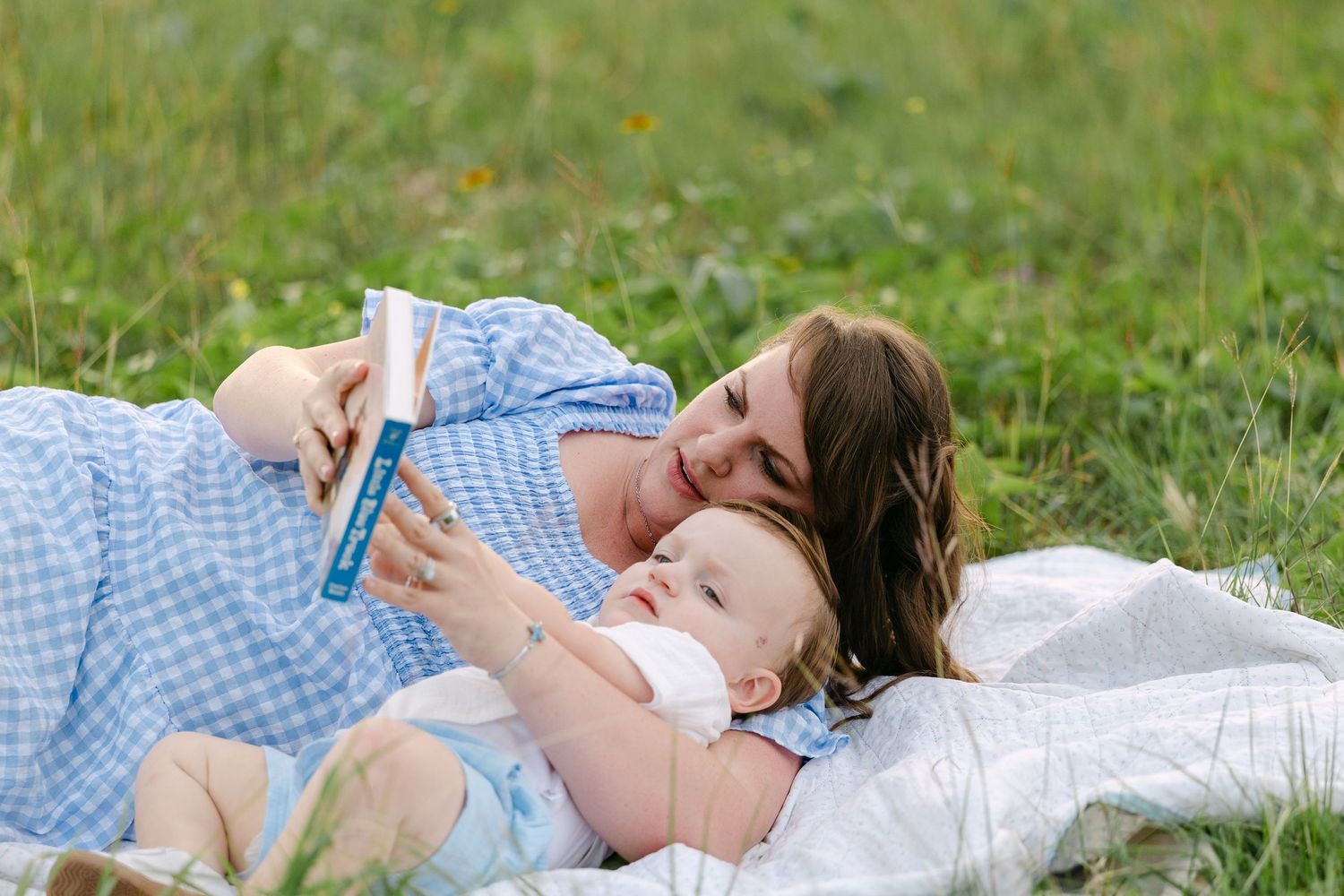 A mother and baby lie on a blanket in green grass, sharing a book together outdoors.