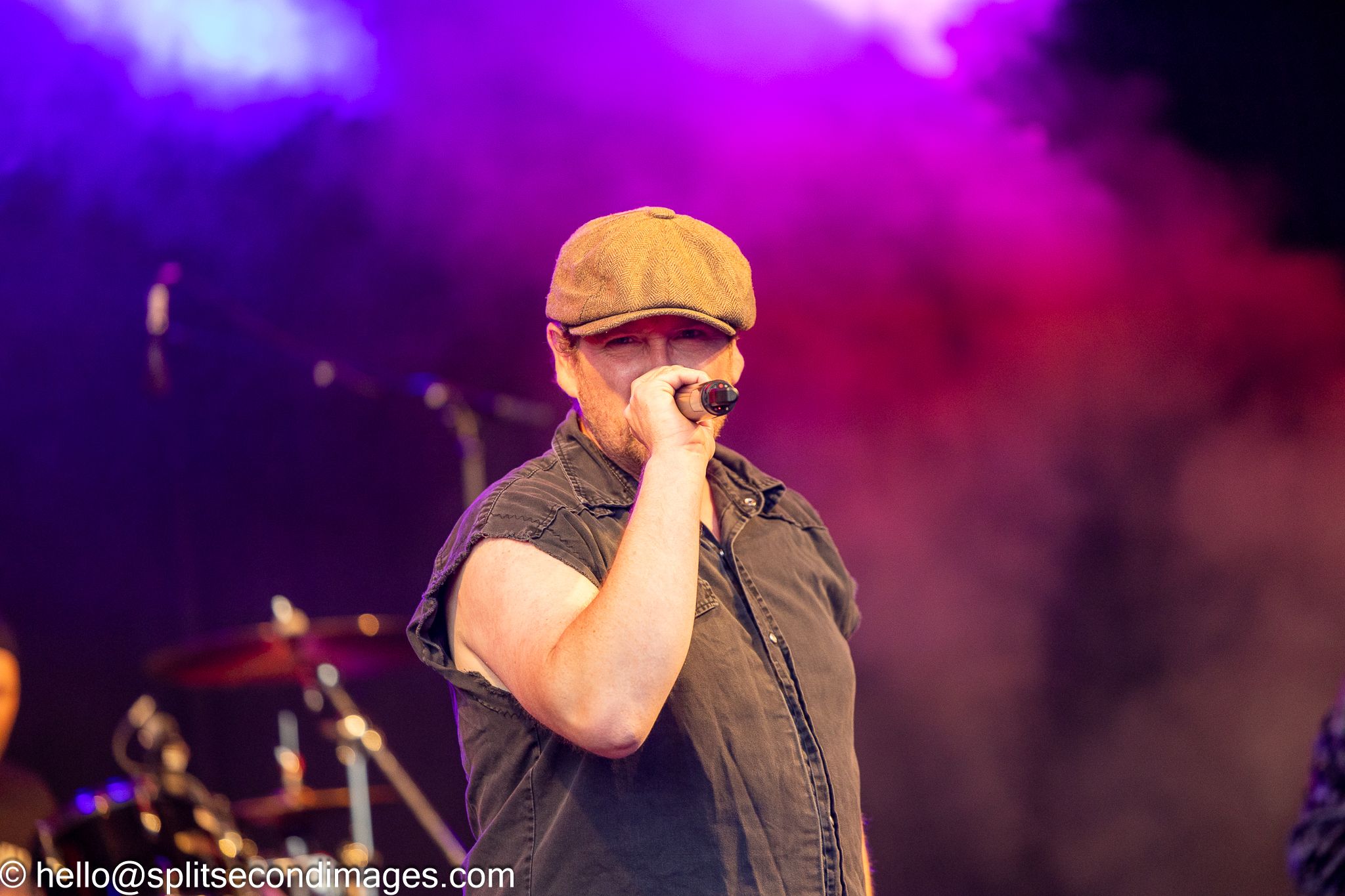 Male vocalist performing with microphone wearing brown cap at outdoor music festival.