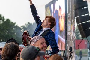 Guitarist crowd surfing while playing guitar at outdoor music festival event.