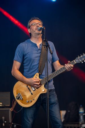 Male guitarist in blue t-shirt singing into microphone while playing a natural wood electric guitar on stage.