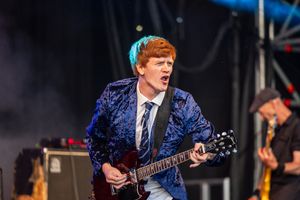 Red-haired guitarist in blue velvet blazer performing energetically on an outdoor festival stage.