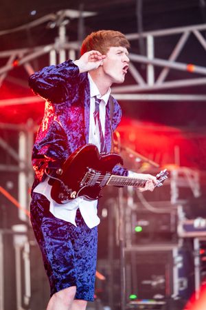 Young male guitarist performing on stage in a blue velvet suit playing a dark electric guitar.