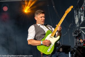 Male guitarist energetically playing green Telecaster electric guitar on outdoor festival stage.