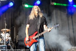 Long-haired male bassist playing red electric bass guitar at outdoor music festival.