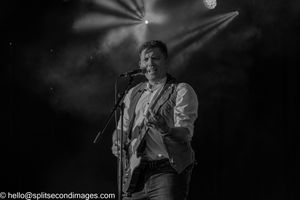 Black and white photo of smiling male guitarist performing on stage with dramatic lighting.