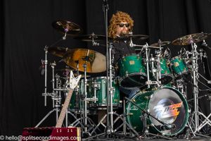 Curly-haired drummer performing on a green drum kit on an outdoor festival stage.