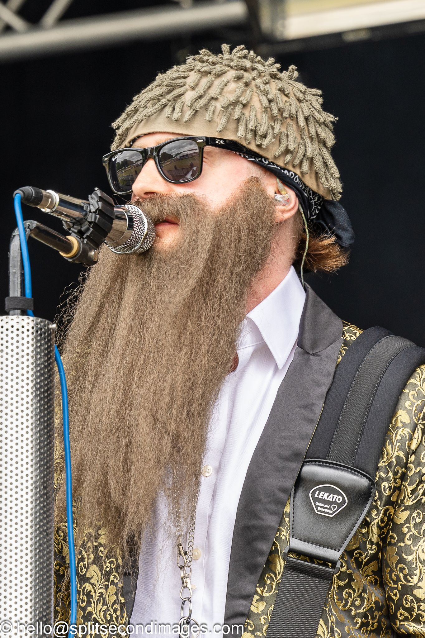 Bearded male musician with long beard wearing sunglasses and dreadlock hat singing into microphone on stage.