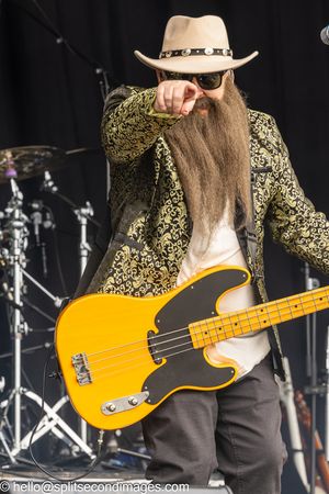 Bearded bassist in cowboy hat and ornate jacket pointing at audience while playing yellow bass guitar.