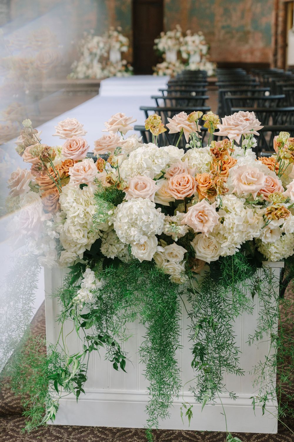 Lush floral arrangement of peach roses and white hydrangeas adorning a white ceremony aisle pedestal.