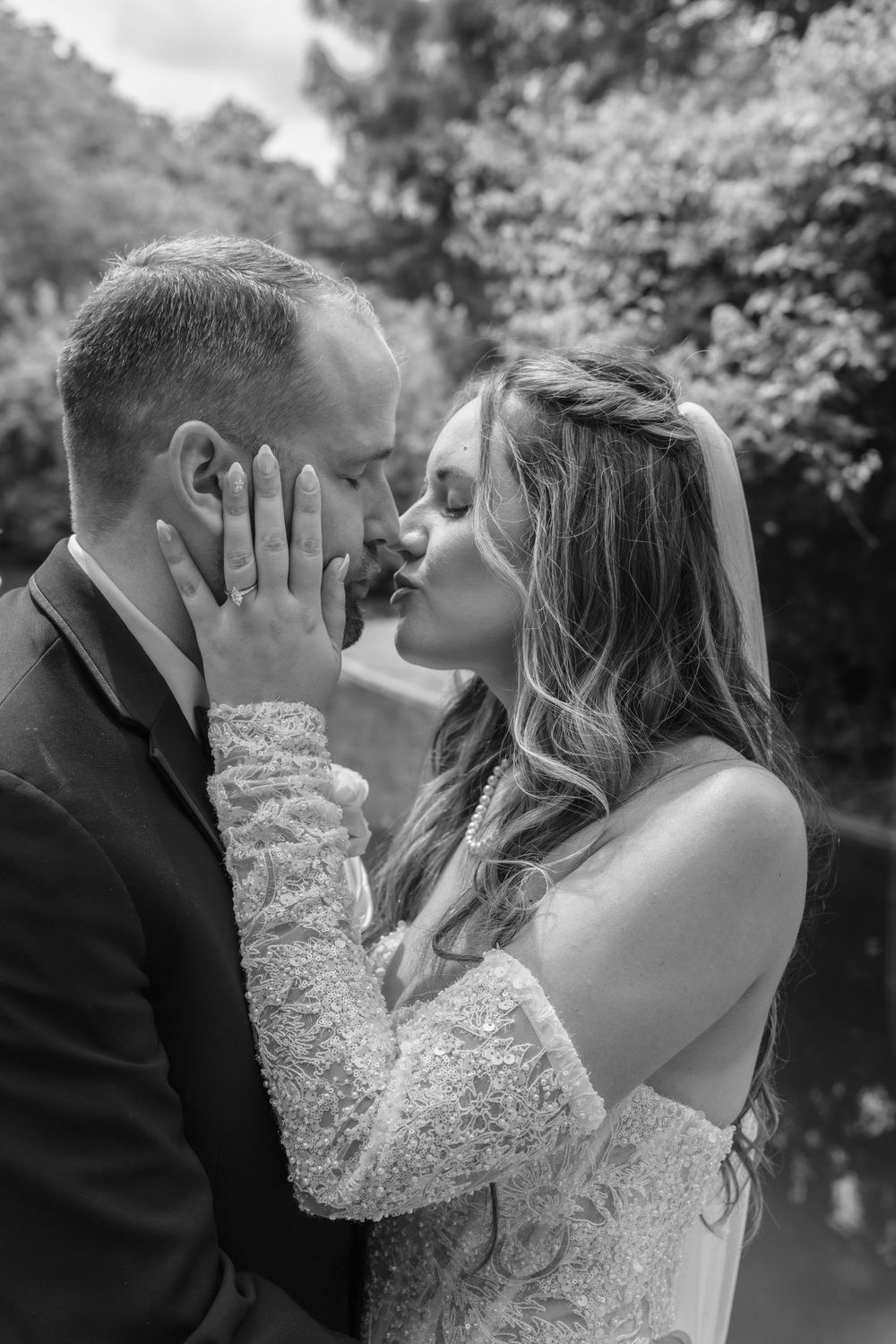 Black and white photo of a bride and groom sharing a tender kiss, bride in lace dress with veil, outdoors.