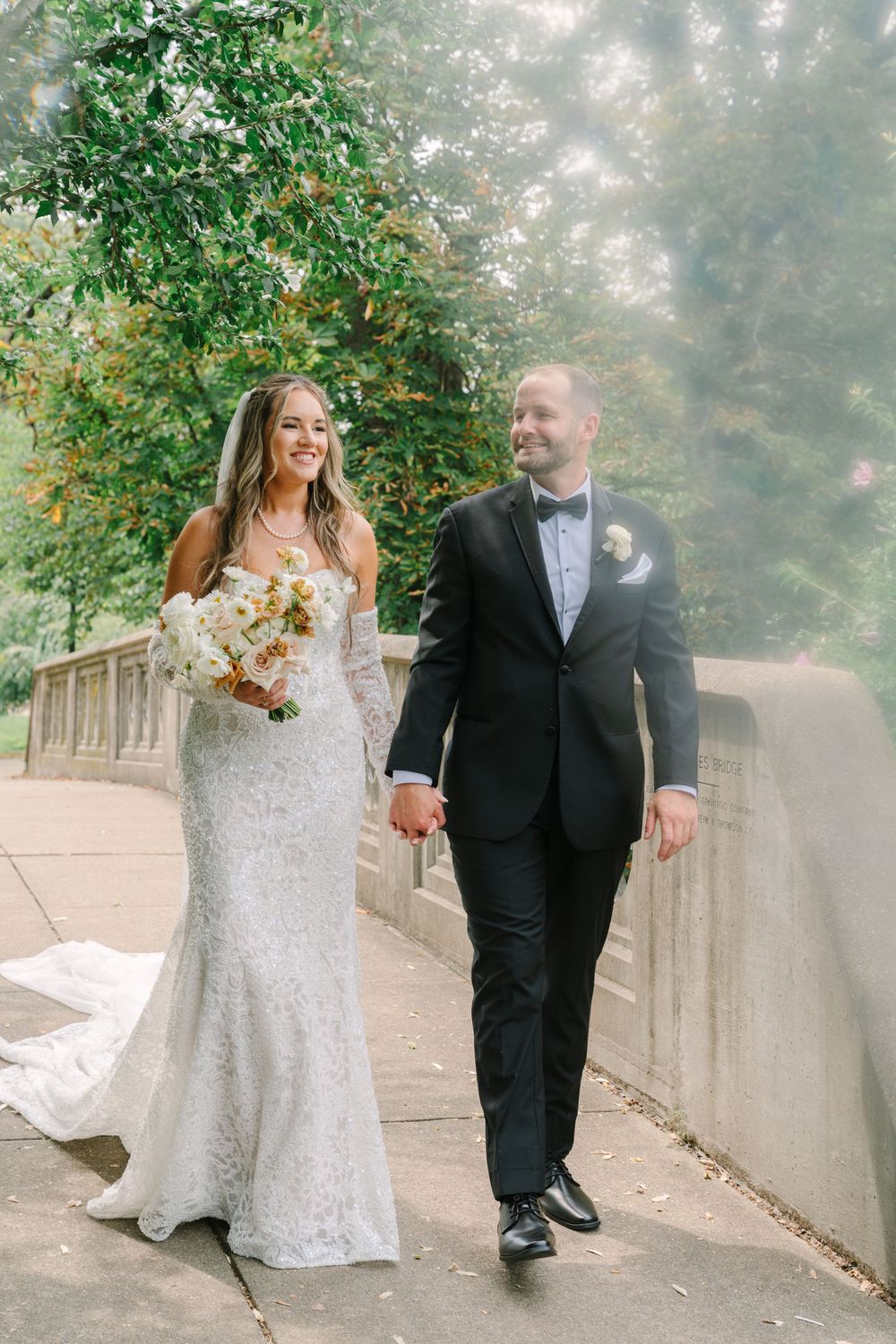 A bride and groom walk hand-in-hand on a bridge surrounded by lush greenery on their wedding day.