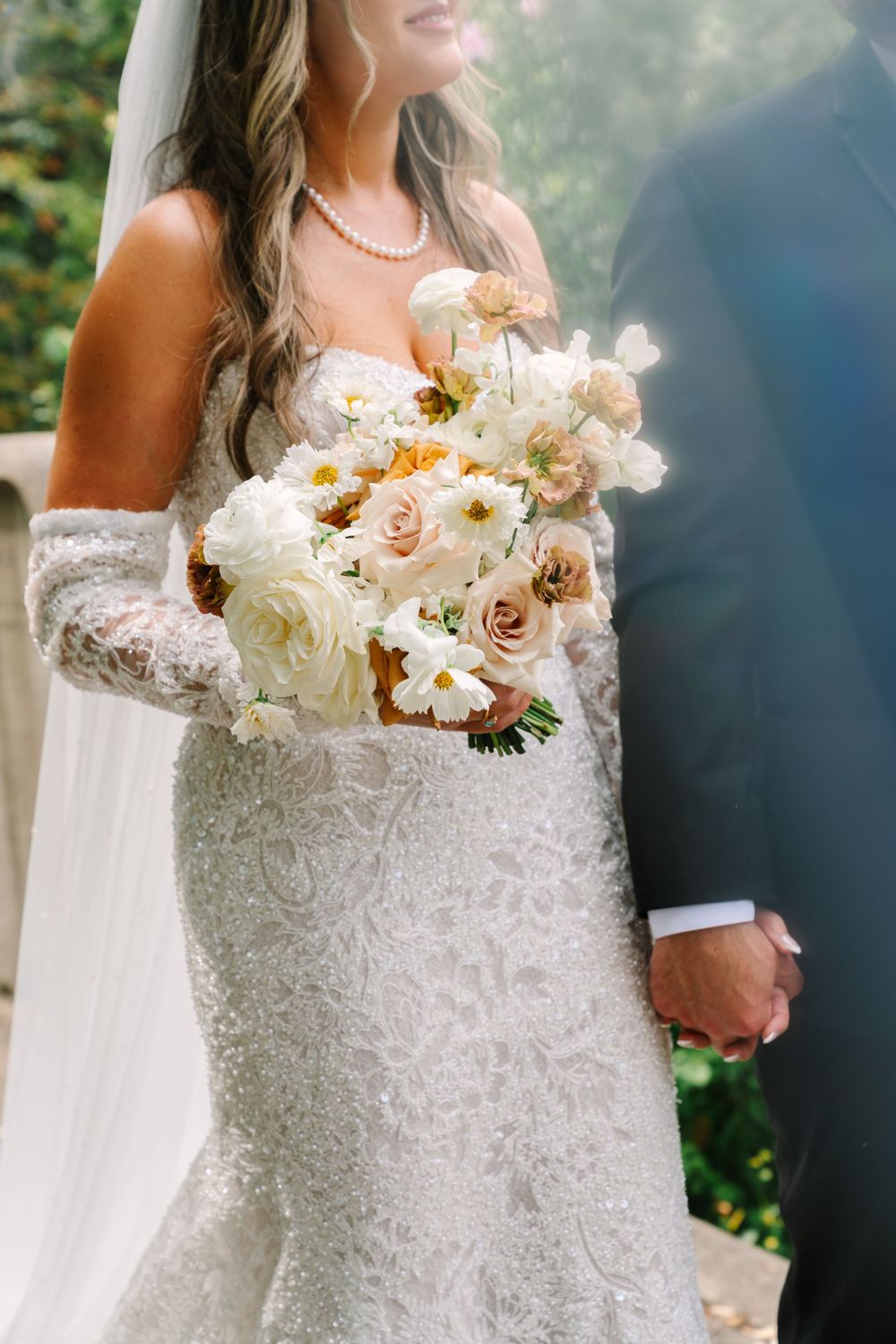 Bride in beaded gown with lace gloves holds white and blush bouquet beside groom outdoors.