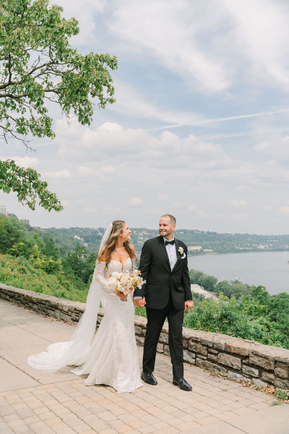 Bride and groom hold hands on a stone terrace overlooking a river, surrounded by lush greenery under a partly cloudy sky.