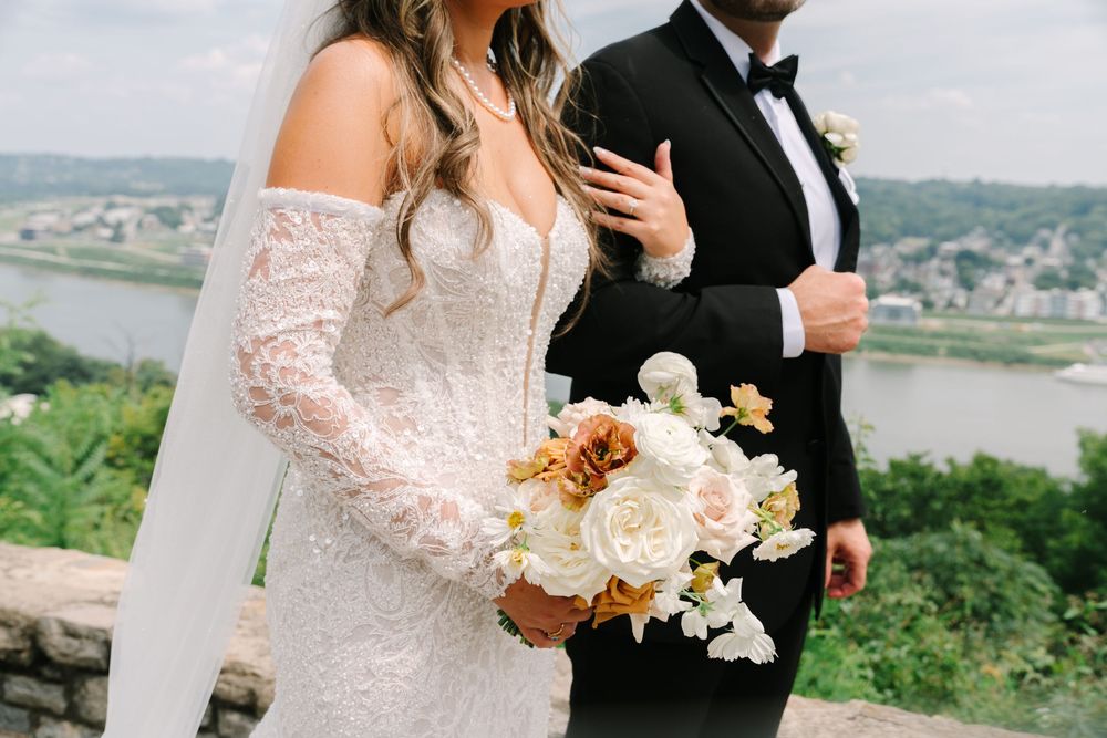 Bride in lace off-shoulder wedding gown holding floral bouquet stands beside groom in black tuxedo with river view backdrop.