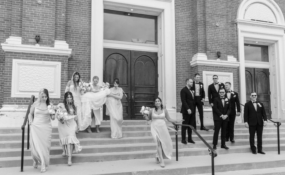 Black and white wedding party photo on church steps, bridesmaids in white gowns and groomsmen in tuxedos.
